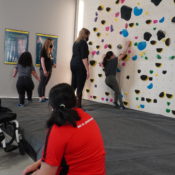ASC MB Athlete Climbing Sideways On The Wall At The Hive While Others Cheer Her On