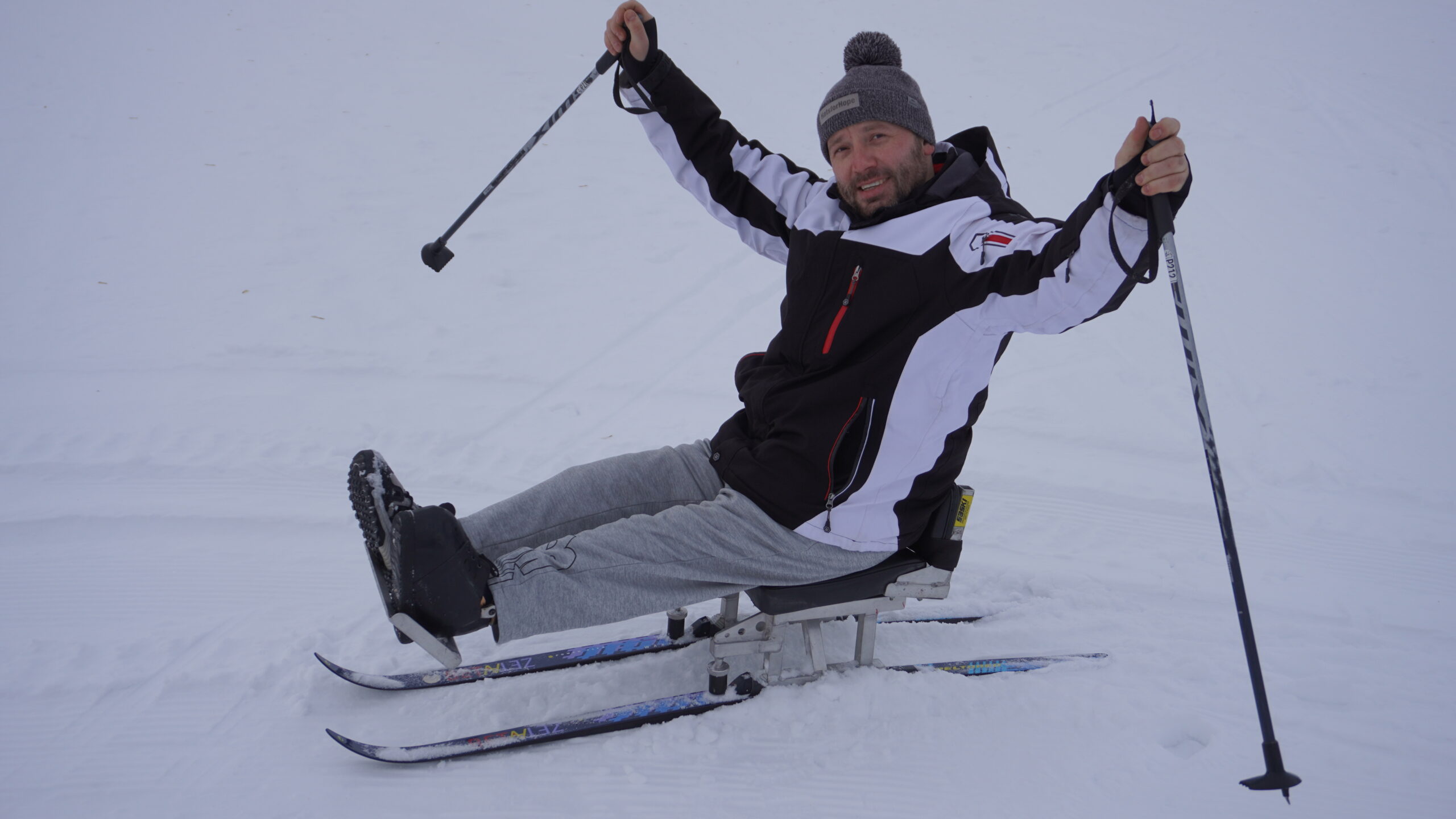 Coach Olex is in a sit ski, with his legs straight out and his arms in the air with ski poles in hand. He has a big grin on his face and welcomes you to join him for a ski!