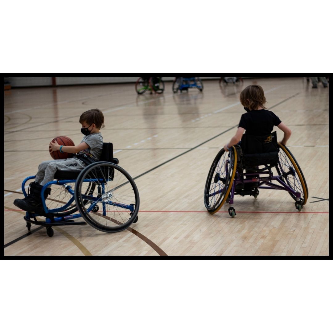 Two young children in wheelchair basketball court chairs, play with a basketball in a gymnasium